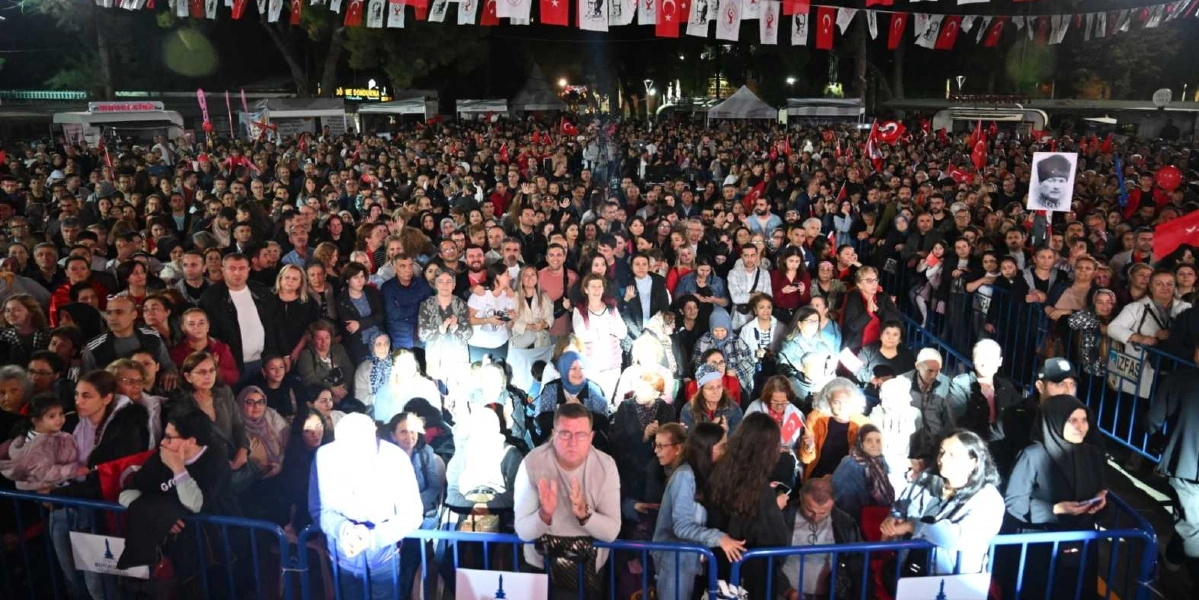 Gece vakti açık havada düzenlenen bir etkinlikte, bariyerlerin arkasında toplanmış büyük bir kalabalık görülüyor. İnsanlar ellerinde Türk bayrakları tutuyor ve birçoğu alkışlıyor. Kalabalığın üzerinde, iplere asılmış çok sayıda Türk bayrağı ve üzerinde Atatürk portresi bulunan flamalar yer alıyor. Sağ tarafta bir kişi, elinde Mustafa Kemal Atatürk'ün fotoğrafının bulunduğu bir pankartı havada tutuyor. Alanın arka planında ise çeşitli stantlar ve ağaçlar seçiliyor.