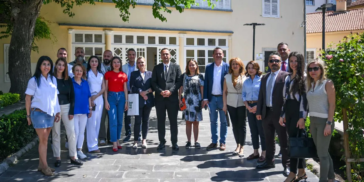 Tarihi bir binanın önündeki bahçede yan yana duran, aralarında Bornova Belediyesi ve Goethe Enstitüsü temsilcilerinin bulunduğu yaklaşık yirmi kişilik grup, objektife gülümseyerek poz veriyor. Gruptaki kişiler günlük ve resmi kıyafetler içinde, bazıları ellerinde hediye çantalarıyla yer alıyor. Arka planda beyaz çerçeveli pencereleri olan sarı renkli bir yapı ve ağaçlar görülüyor.