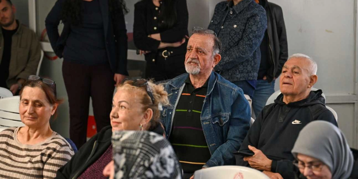 Bir eğitim veya bilgilendirme toplantısında oturan, farklı yaş gruplarından oluşan bir grup insan. Ön planda, çizgili kazak giymiş bir kadın, yanında kısmen görünen başka bir kadın, kot ceketli ve çizgili tişörtlü orta yaşlı bir adam ile siyah Nike sweatshirt giymiş başka bir adam dikkatle dinliyor. Arka planda ise ayakta duran ve oturan diğer katılımcılar yer alıyor.