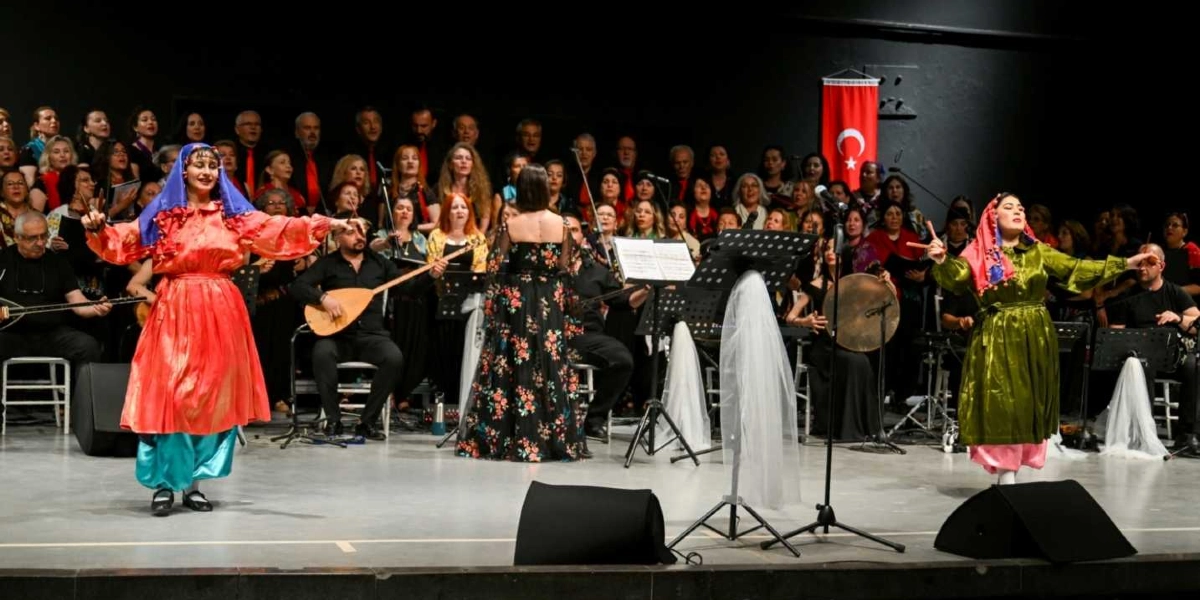 Sahne üzerinde geleneksel kıyafetler giymiş iki kadın dansçı, arkalarında geniş bir koro ve müzisyen grubuyla performans sergiliyor. Soldaki dansçı kırmızı elbise ve mavi başörtüsü, sağdaki dansçı ise yeşil elbise ve kırmızı başörtüsü ile dans ediyor. Arka planda siyah giyimli koro üyeleri, ellerinde enstrümanlar bulunan müzisyenler ve duvarda asılı bir Türk bayrağı yer alıyor. Sahnenin ortasında nota sehpaları ve mikrofonlar bulunuyor.