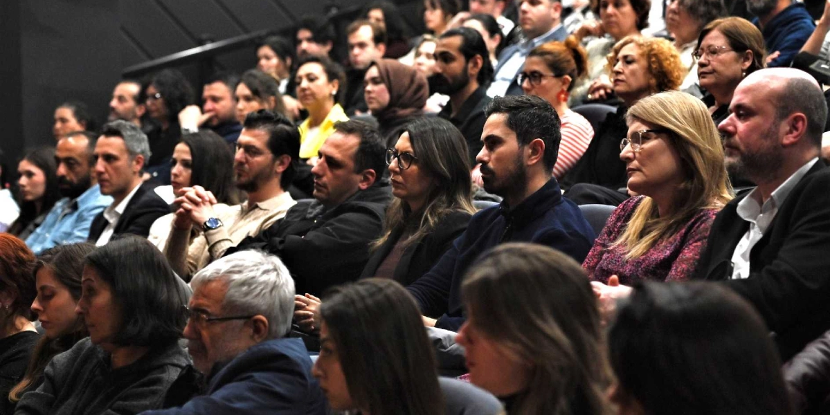 Tiyatro salonunda sahneyi dikkatle izleyen, farklı yaş gruplarından oluşan kalabalık bir seyirci grubu.