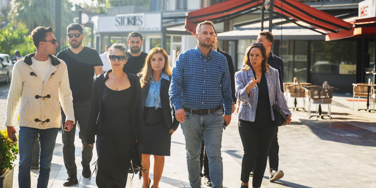 Açık havada, bir cadde üzerinde yürüyen ve aralarında Bornova Belediye Başkanı Ömer Eşmi'nin de bulunduğu bir grup insan, bir noktayı işaret eden bir kadınla birlikte ilerliyor. Gruptakiler günlük ve şık kıyafetler giymiş, çevrelerindeki dükkanların ve restoranların bulunduğu alanda incelemelerde bulunuyorlar.