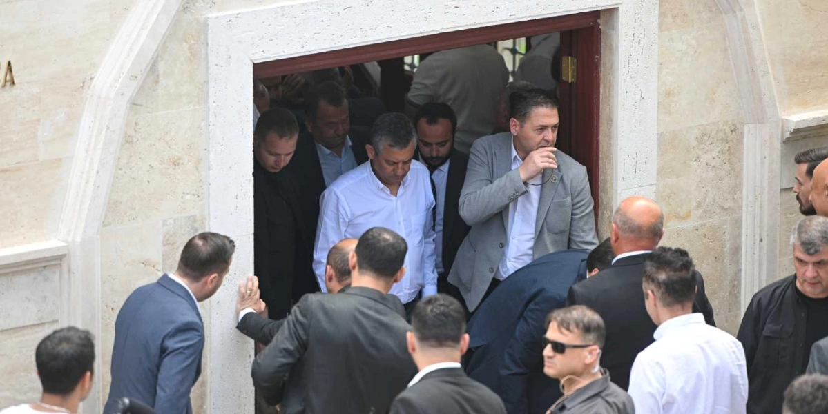 CHP Genel Başkanı Özgür Özel, beyaz gömlekli haliyle bir binanın giriş kapısından dışarıya doğru çıkarken, çevresinde takım elbiseli korumalar ve yetkililer bulunuyor.