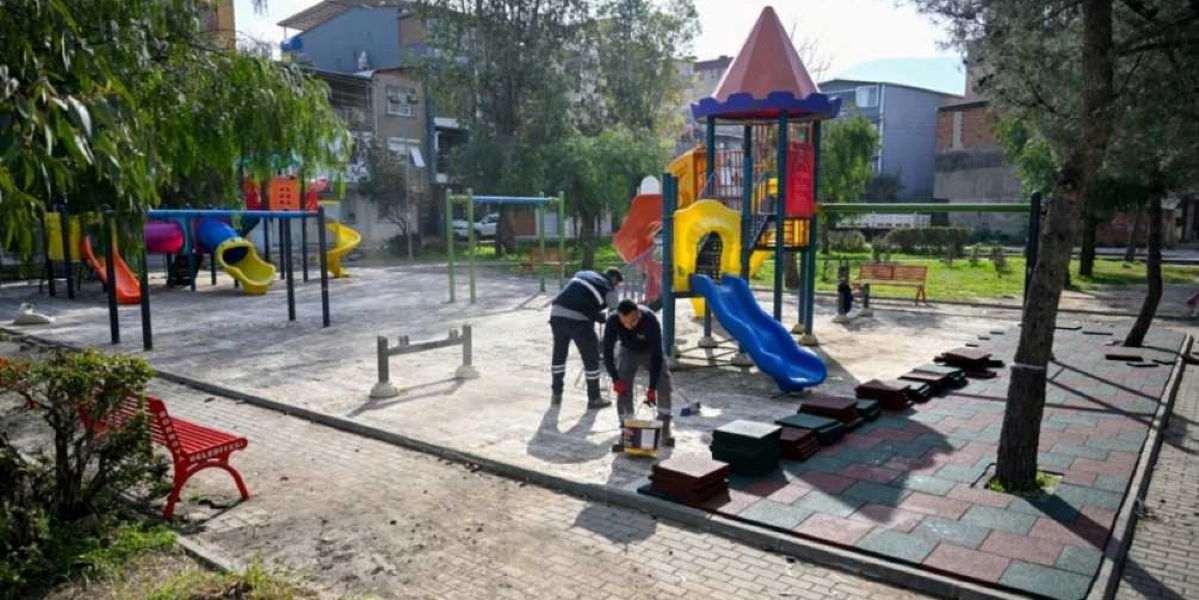 Bornova Belediyesi ekipleri, bir çocuk parkında zemin düzenleme ve bakım çalışması yapıyor. Parktaki iki işçi, oyun gruplarının yanındaki alana kauçuk zemin döşüyor. Parkta renkli kaydıraklar, salıncaklar ve üzerinde Bornova Belediyesi yazan kırmızı bir bank bulunuyor.