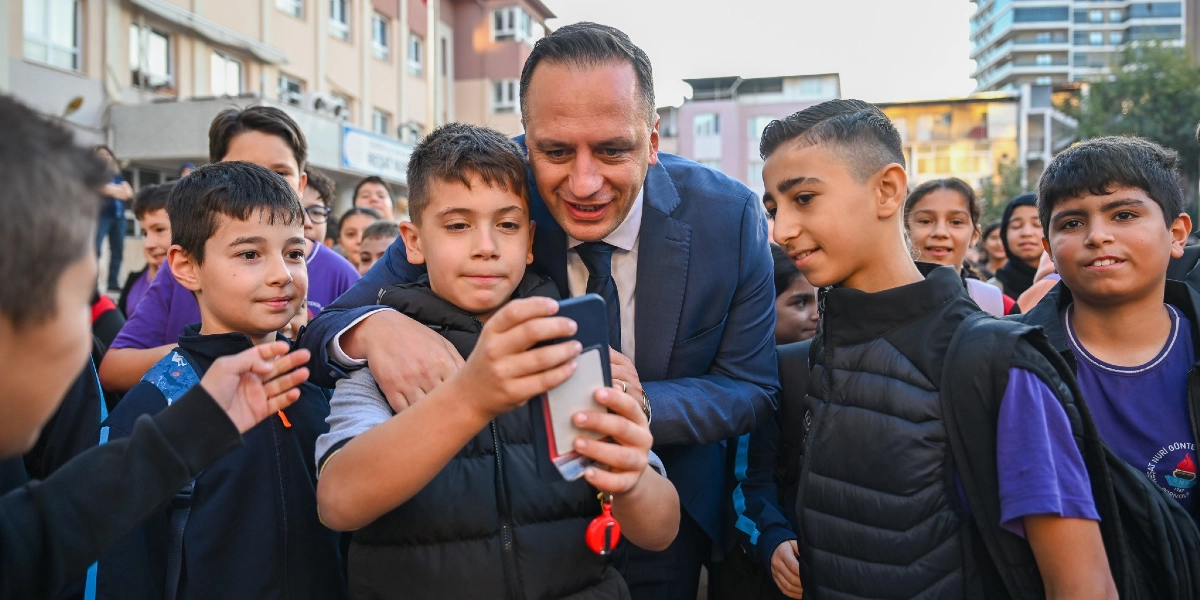 Lacivert takım elbiseli bir erkek, okul bahçesinde etrafını saran ilkokul çağındaki öğrencilerle birlikte gülümseyerek bir akıllı telefona bakıyor. Öğrencilerden biri telefonu tutarken, takım elbiseli kişi elini çocuğun omzuna atmış durumda. Arka planda okul binası ve diğer öğrenciler görülüyor.