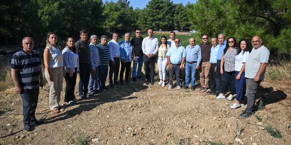 Açık havada, ağaçlık bir alanın önündeki toprak arazide yan yana duran, çoğunluğu erkeklerden oluşan yaklaşık yirmi kişilik bir grup insan. Günlük kıyafetler içindeki grup, kameraya bakarak poz veriyor.