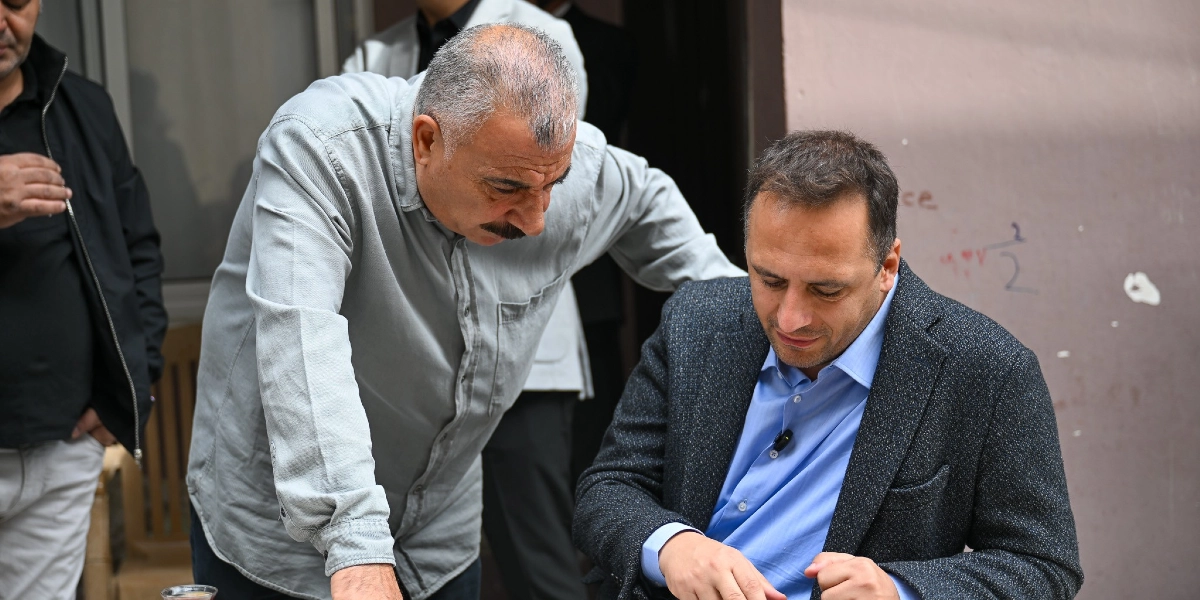 Gri gömlekli, bıyıklı orta yaşlı bir adam, masada oturan mavi gömlekli ve koyu renk ceketli bir adamın yanına eğilmiş, dikkatle bir şeye bakıyorlar. Arka planda başka insanlar da bulunuyor.