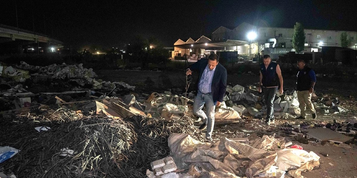 Gece vakti açık bir arazide bir araya gelen üç kişi, yığılmış molozların ve metal atıkların arasında incelemelerde bulunuyor. Lacivert ceketli bir adam elindeki metal bir parçayı incelerken, arkasında belediye üniformalı iki kişi onu takip ediyor. Arka planda ise aydınlatılmış bir fabrika veya depo binası ile bir üst geçit silüeti görülüyor.