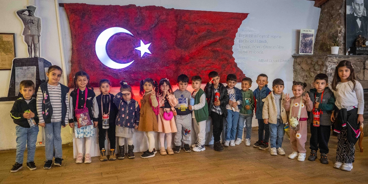 Belkahve'deki bir müze ortamında, arkalarında büyük, dokulu kırmızı bir zemin üzerine yerleştirilmiş ışıklı Türk bayrağı bulunan yaklaşık yirmi çocuk yan yana dizilmiş poz veriyor. Çocukların solunda Mustafa Kemal Atatürk'ün asker kıyafetli bir maket figürü, sağında ise duvarda Atatürk'ün portresi ve Mithat Cemal Kuntay'ın 