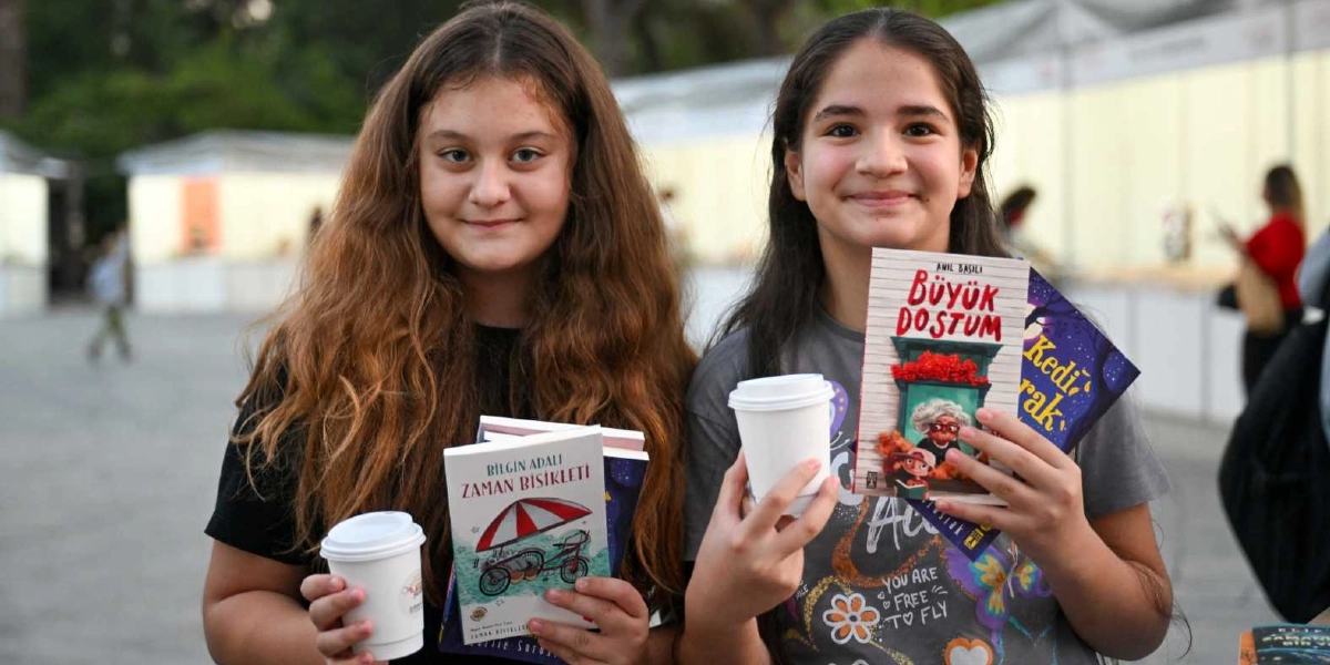 Açık havada düzenlenen bir kitap fuarında, ellerinde karton bardaklarla içeceklerini tutan ve gülümserek kameraya bakan iki genç kız. Soldaki kız elinde Bilgin Adalı'nın Zaman Bisikleti kitabını, sağdaki kız ise Anıl Basılı'nın Büyük Dostum kitabını tutuyor. Arka planda fuar stantları ve yürüyen insanlar görülüyor.