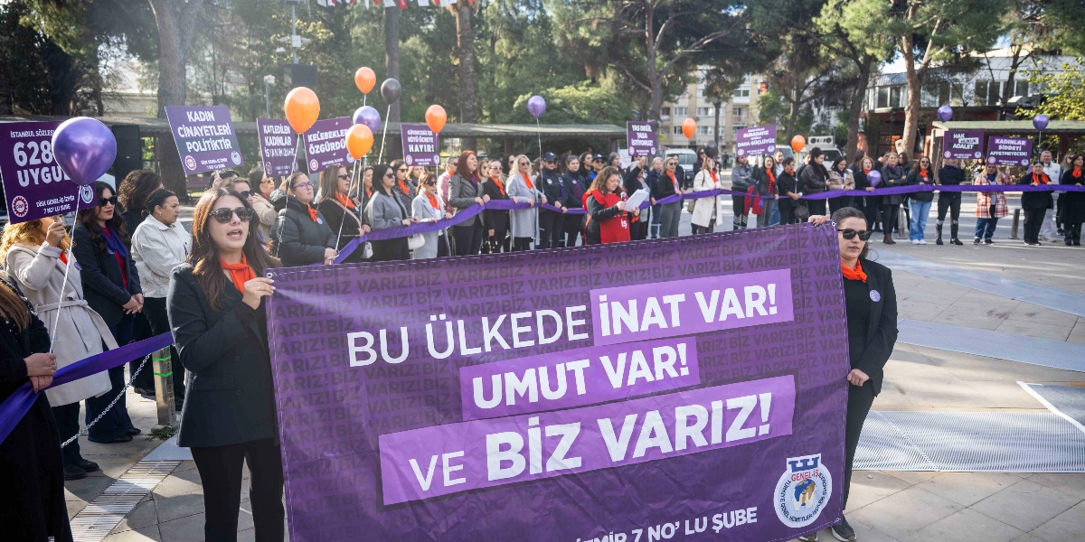 Açık havada, ağaçlık bir alanda bir araya gelmiş kadınlar grubu, ellerinde mor renkli pankartlar ve balonlarla 25 Kasım Kadına Yönelik Şiddete Karşı Uluslararası Mücadele Günü kapsamında bir eylem gerçekleştiriyor. Grubun önünde tutulan büyük mor pankartın üzerinde beyaz harflerle 