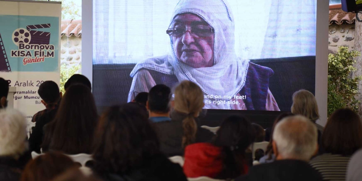 Bornova Kısa Film Günleri etkinliğinde, izleyiciler beyaz perdede başörtülü ve gözlüklü yaşlı bir kadının yer aldığı bir belgesel veya film kesitini izliyor. Ekranın sol tarafında etkinliğin logosunun ve tarihlerinin bulunduğu dikey bir afiş yer alıyor.