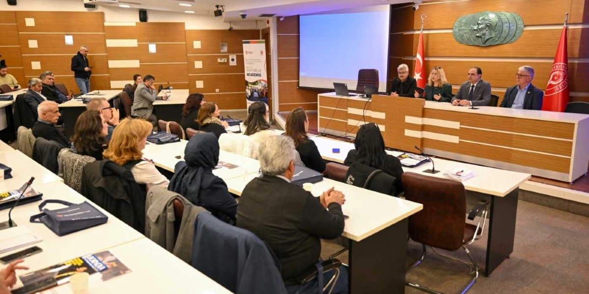 Modern bir toplantı salonunda, ön taraftaki masada oturan dört konuşmacı ve onları dinleyen katılımcıların bulunduğu bir etkinlik. Salonun ön kısmında, üzerinde Türk bayrağı ve Bornova Belediyesi flaması bulunan ahşap bir kürsü masası yer alıyor. Arka duvarda Mustafa Kemal Atatürk'ün kabartma portresi ve boş bir projeksiyon perdesi bulunuyor. Katılımcılar sıralı masalarda oturuyor, yan tarafta ise üzerinde 