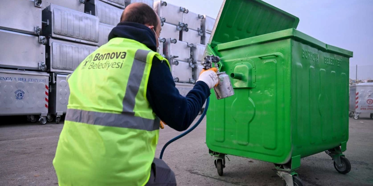 Bornova Belediyesi logolu fosforlu sarı iş yeleği giyen bir belediye çalışanı, elindeki boya tabancasıyla metal bir çöp konteynerini yeşile boyuyor. Arka planda ise henüz boyanmamış, üst üste dizilmiş çok sayıda metal çöp konteyneri bulunuyor.
