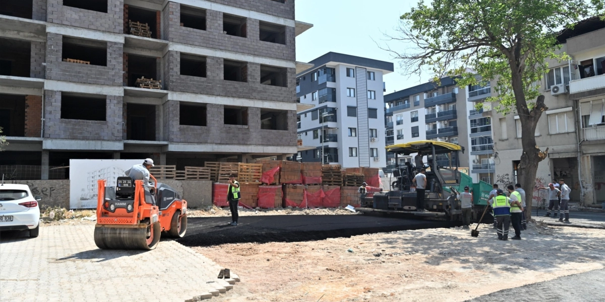 Bornova'da devam eden bir inşaat alanında, turuncu bir silindir ve asfalt serme makinesi kullanılarak yol çalışması yapılıyor. İşçiler asfalt serme işlemini yürütürken, arka planda kaba inşaat halindeki bir bina ve tamamlanmış modern apartmanlar yer alıyor.