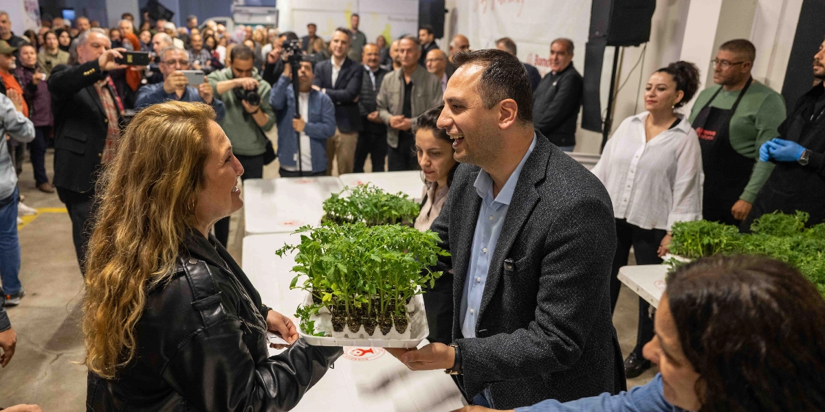 Bornova Belediye Başkanı Mustafa İduğ, kapalı bir alanda düzenlenen etkinlikte bir kadın üreticiye fide tepsisi takdim ediyor. İkili gülümseyerek birbirine bakarken, masanın üzerinde başka fide tepsileri de bulunuyor. Arka planda ise etkinliği takip eden kalabalık bir grup insan, basın mensupları ve belediye çalışanları yer alıyor.