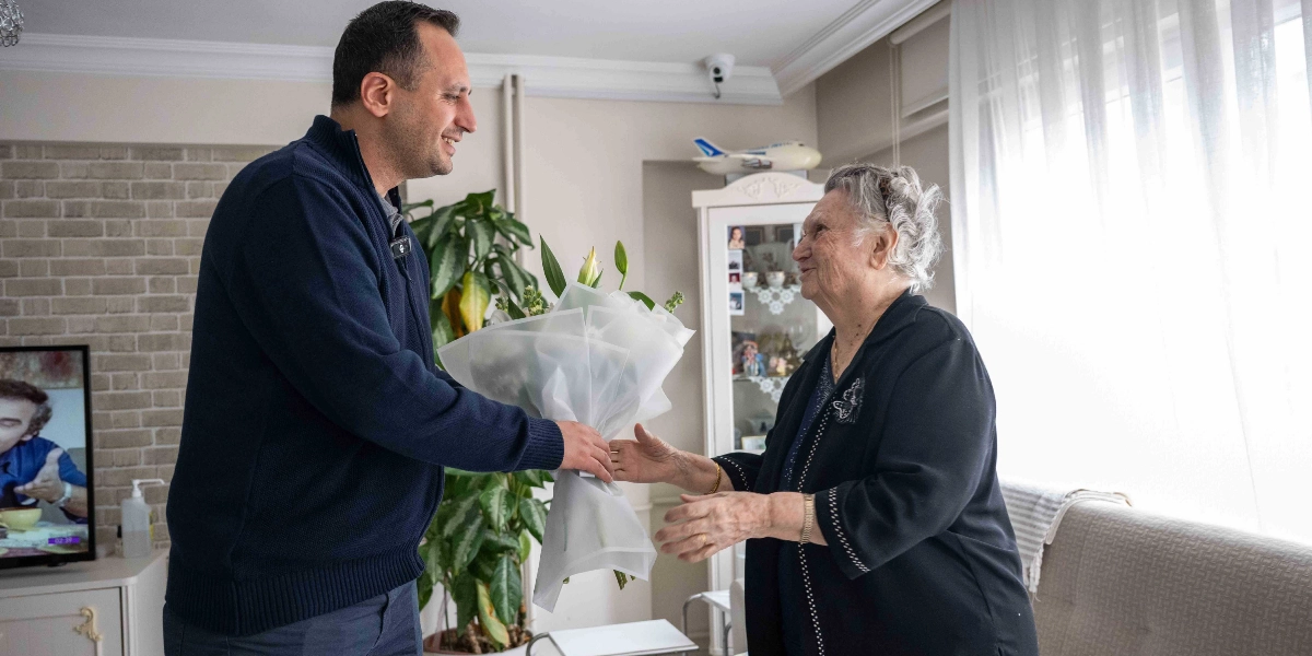 Koyu renkli bir kazak giymiş genç bir adam, ev ortamında yaşlı bir kadına beyaz ambalajlı bir çiçek buketi uzatıyor. Yaşlı kadın, üzerinde siyah bir hırka ile gülümseyerek çiçeği kabul ediyor. Arka planda bir bitki, vitrin ve pencere önünde tül perde yer alıyor.
