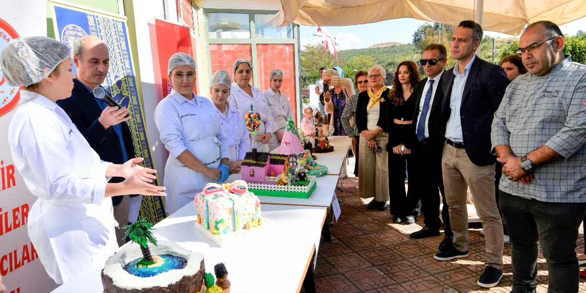 Bornova'da düzenlenen bir pastacılık etkinliğinde, beyaz önlüklü ve boneli kursiyerler hazırladıkları tematik pastaları sergiliyor. Ön planda ada temalı, hediye paketi görünümlü ve masalsı ev figürlü pastalar yer alıyor. Bir eğitmen mikrofonla konuşurken, yanındaki kursiyerler projelerini anlatıyor. Etkinliği takip eden bir grup davetli ise masanın diğer tarafında durarak çalışmaları inceliyor.