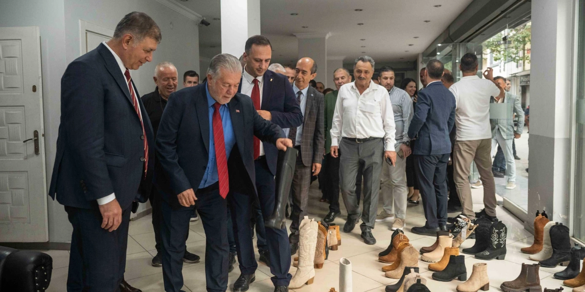 Takım elbiseli bir grup erkek, bir mağaza içerisinde sergilenen çeşitli bot ve ayakkabı modellerini inceliyor. Grubun önünde yer alan, lacivert ceketli ve kırmızı kravatlı bir kişi, elinde tuttuğu siyah bir çizmeye dikkatle bakıyor. Çevresindeki diğer kişiler ise bu incelemeyi takip ediyor.