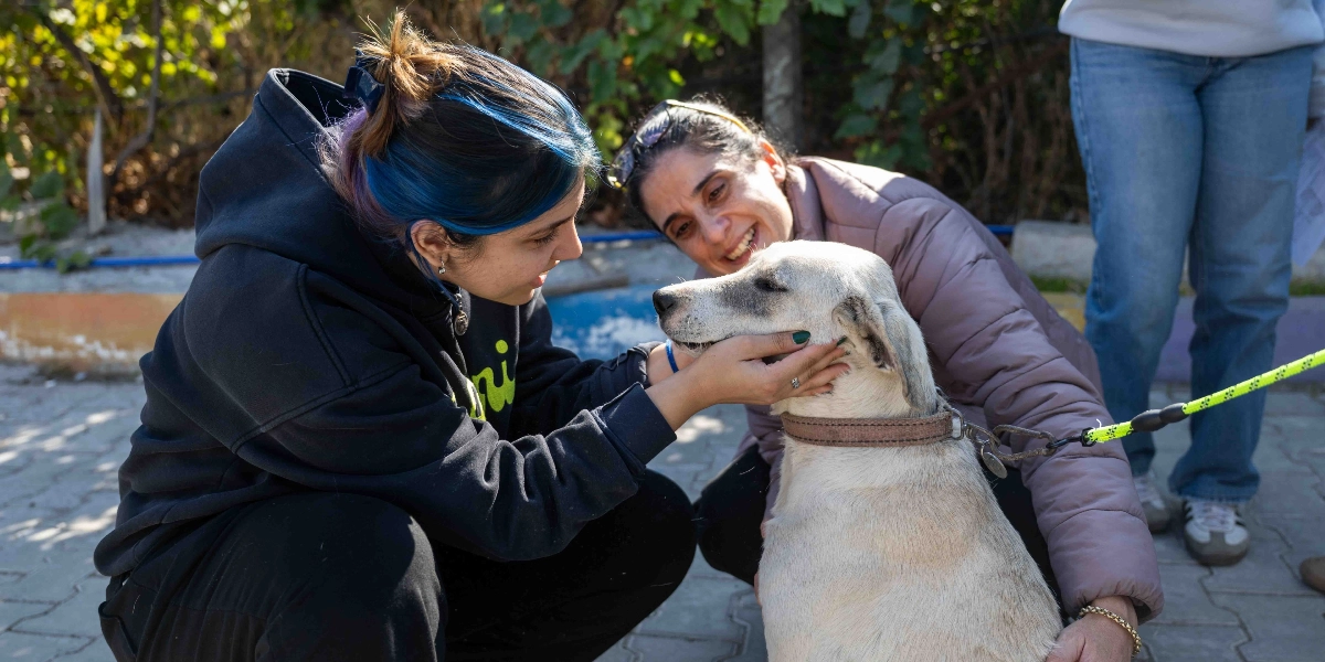 Mavi saçlı genç bir kadın ve yanında duran orta yaşlı bir kadın, dışarıda tasmalı, açık renkli bir köpeği sevgiyle okşuyor. Genç kadın çömelmiş bir şekilde köpeğin yüzünü elleriyle tutarken, yanındaki kadın da gülümseyerek onlara eşlik ediyor.