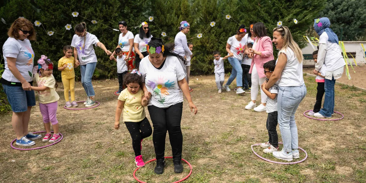Açık havada, toprak bir alanda Anneler Günü etkinliği kapsamında bir araya gelen anneler ve çocuklar, yere konulan hulahopların içinde oyunlar oynuyor. Çoğu beyaz tişört giymiş olan katılımcıların bir kısmı başlarına çiçekli taçlar takmış, neşeli bir şekilde birbirleriyle etkileşim kuruyor. Arka planda yeşil ağaçlar ve çiçek süslemeleri yer alıyor.