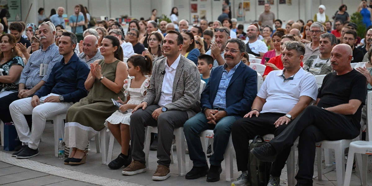 Açık havada düzenlenen bir etkinlikte, beyaz plastik sandalyelerde yan yana oturarak sahneyi izleyen kalabalık bir grup insan. Ön sırada, aralarında Bornova Belediye Başkanı Mustafa İduğ'un da bulunduğu çeşitli yaşlardan kadın, erkek ve bir kız çocuğu yer alıyor. Katılımcıların çoğu dikkatle karşıya bakarken, bazıları alkışlıyor ve genel atmosfer oldukça sakin ve odaklanmış bir şekilde ilerliyor. Arka planda ise etkinliği takip eden daha geniş bir izleyici kitlesi bulunuyor.