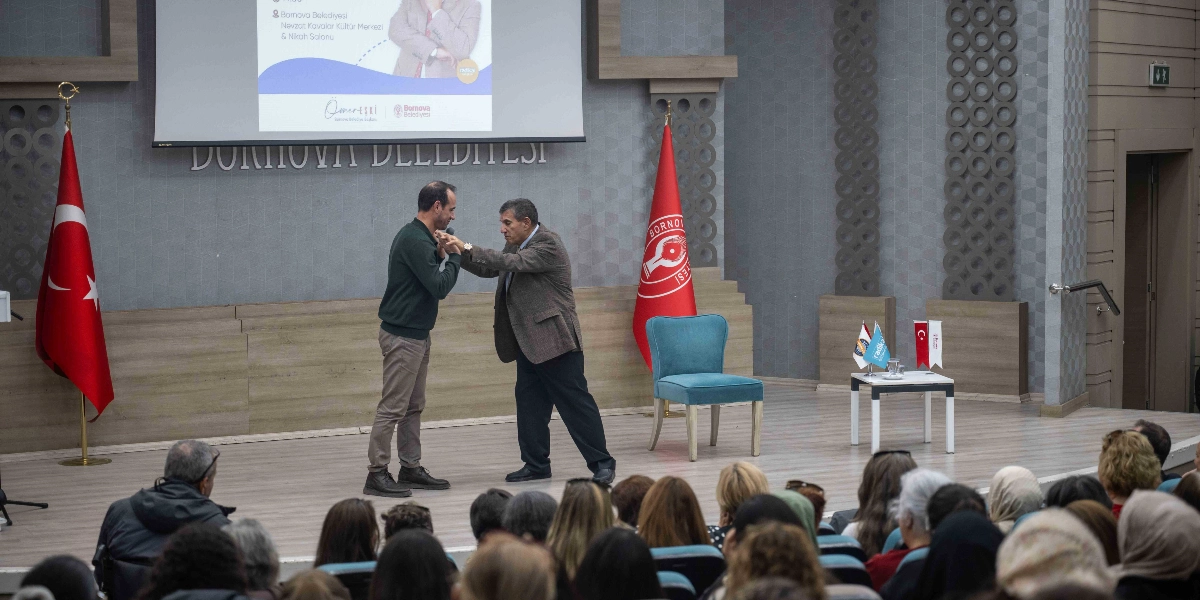Bornova Belediyesi Nevzat Kavalar Kültür Merkezi'nde düzenlenen bir etkinlikte, sahnede konuşmacı olan iki erkek birbirlerinin ellerini tutarak samimi bir şekilde selamlaşıyor. Sahnenin arka planında üzerinde 
