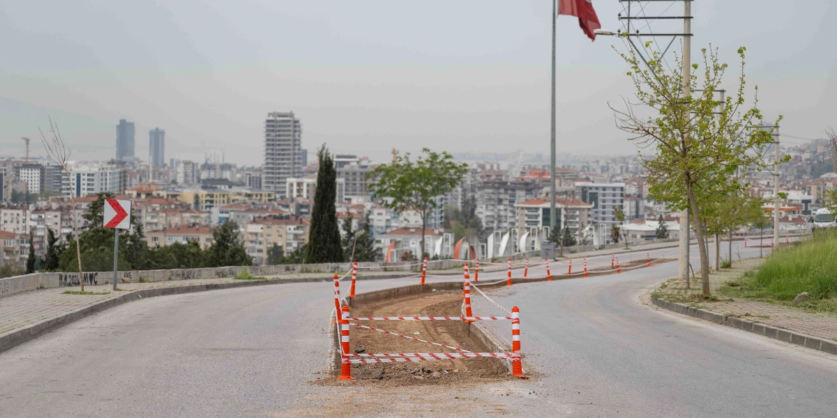 Bornova'da bir yol ayrımında, kırmızı beyaz plastik dubalar ve güvenlik şeritleriyle çevrilmiş, kazı çalışması yapılan toprak bir alan görülüyor. Arka planda ise şehir yerleşimi ve yüksek binalar yer alıyor.