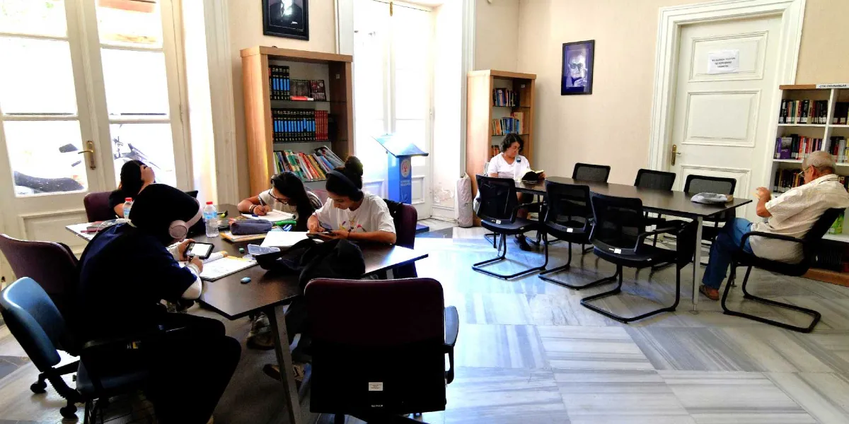 Aydınlık ve geniş bir kütüphane salonunda, masalarda ders çalışan veya kitap okuyan çeşitli yaşlardan insanlar bulunuyor. Ön plandaki masada kulaklık takan ve not alan öğrenciler yer alırken, arka planda başka bir masada kitap okuyan bir kadın ve tek başına oturan yaşlı bir adam görülüyor. Salonun duvarlarında kitap rafları, portreler ve Bornova Belediyesi logosunu taşıyan bir kürsü yer alıyor. Zemin mermer kaplı olup, ortam sakin ve çalışma odaklı bir atmosfer sunuyor.