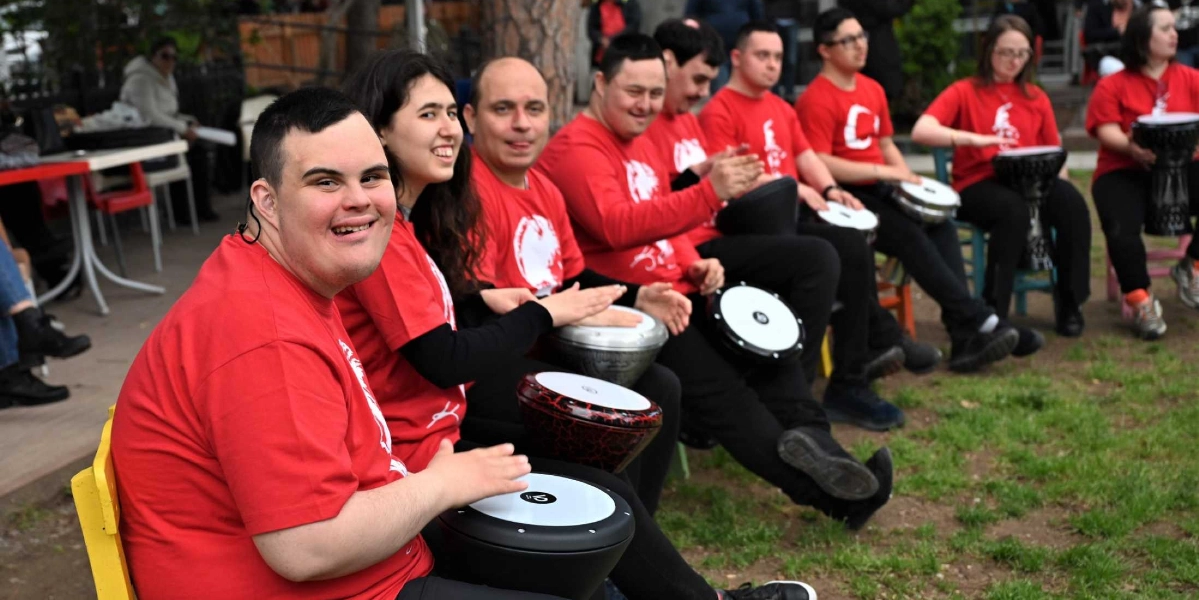 Kırmızı tişörtler giymiş, Down sendromlu bireylerden oluşan bir grup, açık havada çimlerin üzerinde oturarak ellerindeki darbuka ve benzeri vurmalı çalgıları çalıyor. Gruptaki herkes neşeli bir ifadeyle müziğe eşlik ederken, ön planda oturan genç bir erkek gülümseyerek kameraya bakıyor.