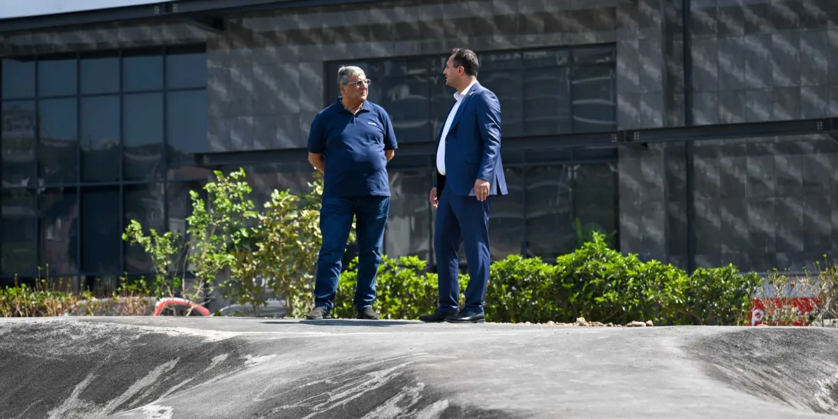 Modern bir binanın önünde, asfalt zeminli engebeli bir parkur üzerinde duran iki erkekten biri lacivert polo yaka tişört ve kot pantolon, diğeri ise lacivert takım elbise giymiş şekilde birbirleriyle konuşuyor.