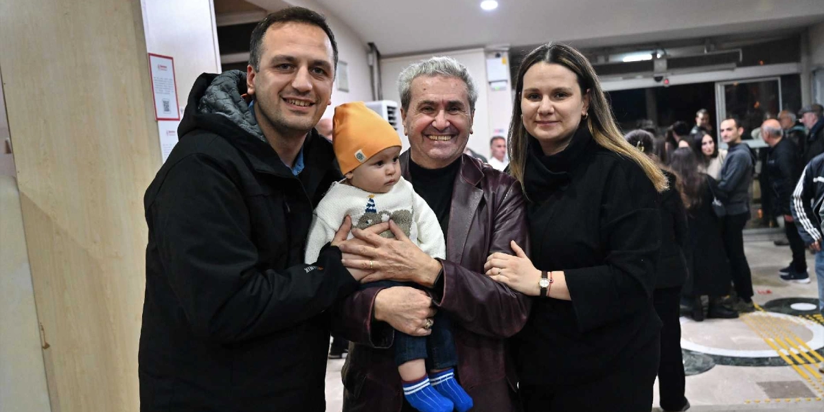 Tiyatro binasının fuaye alanında, kucağında turuncu bereli bir bebek tutan orta yaşlı bir adam, yanında duran genç bir çiftle birlikte gülümseyerek poz veriyor. Adam koyu renk deri bir ceket giyerken, yanındaki erkek siyah bir mont, kadın ise siyah bir kazak giyiyor. Arka planda tiyatro çıkışında bekleyen diğer insanlar ve binanın giriş kapısı görülüyor.