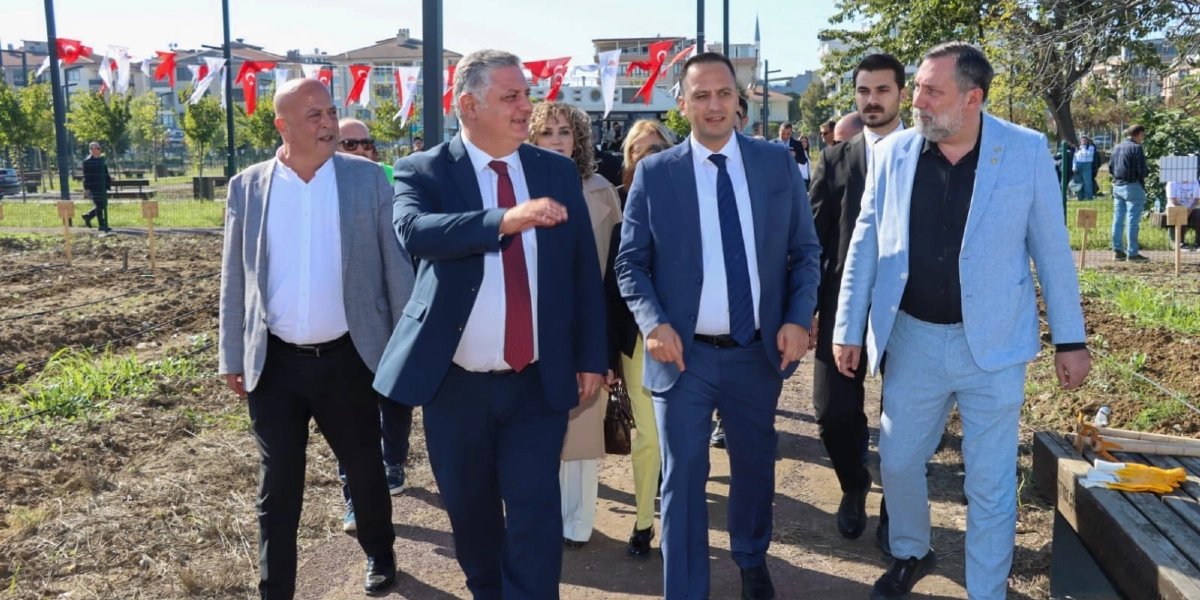 Açık havada, arkada Türk bayraklarıyla süslenmiş bir alanda yürüyen, takım elbiseli bir grup erkek ve kadın yetkili. Ön planda, lacivert takım elbiseli ve kırmızı kravatlı bir kişi, yanındaki mavi takım elbiseli kişiye bir şeyler anlatarak işaret ediyor. Grubun geri kalanında gri ve açık mavi ceketli erkekler ile sarı pantolonlu bir kadın yer alıyor. Arka planda ise ağaçlar, binalar ve toprak bir arazi görülüyor.