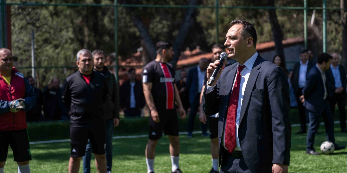 Lacivert takım elbiseli ve kırmızı kravatlı Başkan Eşki, bir halı sahada elinde mikrofonla konuşma yapıyor. Arka planda futbol forması giymiş oyuncular ve takım elbiseli izleyiciler yer alıyor. Sahada bir futbol topu duruyor ve çevresi tel örgülerle çevrili.