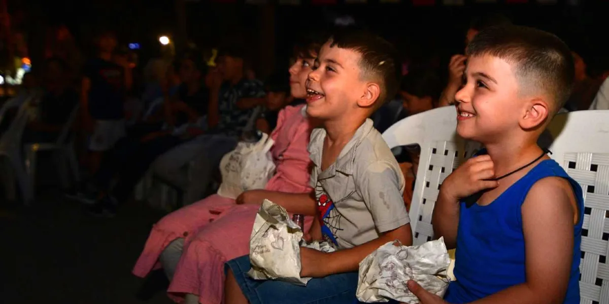 Gece vakti açık hava sinemasında yan yana oturan, ellerinde patlamış mısır paketleri bulunan ve neşeyle ekrana bakan çocuklar.