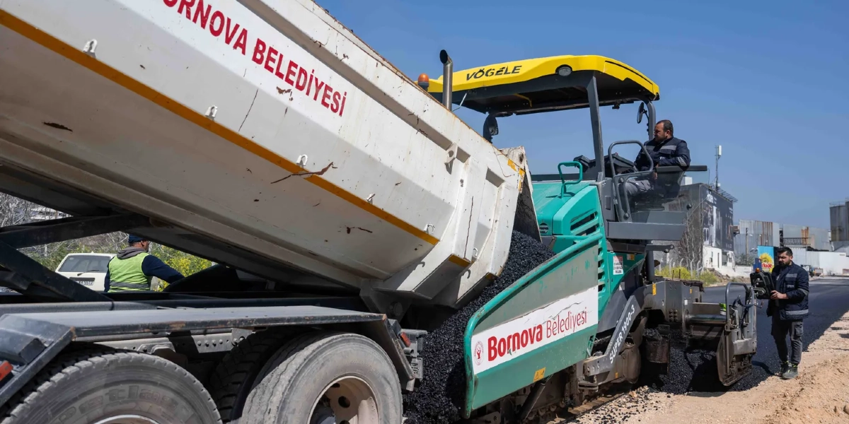 Bornova Belediyesi'ne ait beyaz bir damperli kamyon, asfalt serme makinesine sıcak asfalt boşaltıyor. Yeşil renkli Vögele marka asfalt serici makinesinin üzerinde bir operatör bulunurken, bir işçi de makinenin yanında elindeki kumanda ile çalışmaları yönetiyor. Çalışma alanı açık havada, güneşli bir günde gerçekleşiyor.