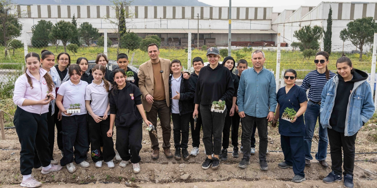Açık havada, toprak bir alanda bir araya gelmiş bir grup öğrenci, öğretmen ve yetkili, ellerinde küçük fide viyolleri tutarak poz veriyor. Arka planda tel örgülerle çevrili bir arazi ve büyük bir sanayi yapısı bulunuyor. Katılımcıların bir kısmı ellerindeki fideleri dikmeye hazırlanırken, bir kısmı gülümsüyor; ortamda kolektif bir tarım çalışması havası hakim.