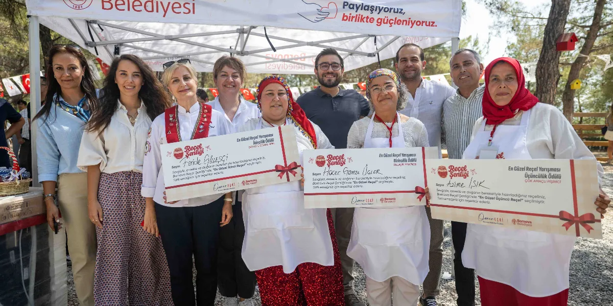 Yakaköy'de düzenlenen Reçel Şenliği'nde, En Güzel Reçel Yarışması'nda dereceye giren üç kadın katılımcı, ellerinde ödül çekleriyle birlikte belediye yetkilileriyle yan yana durarak poz veriyor. Önde duran üç kadın, üzerinde isimlerinin ve kazandıkları derecelerin yazılı olduğu büyük ödül belgelerini tutuyor. Arka planda Bornova Belediyesi logolu beyaz bir çadır ve ağaçlık bir alan bulunuyor.