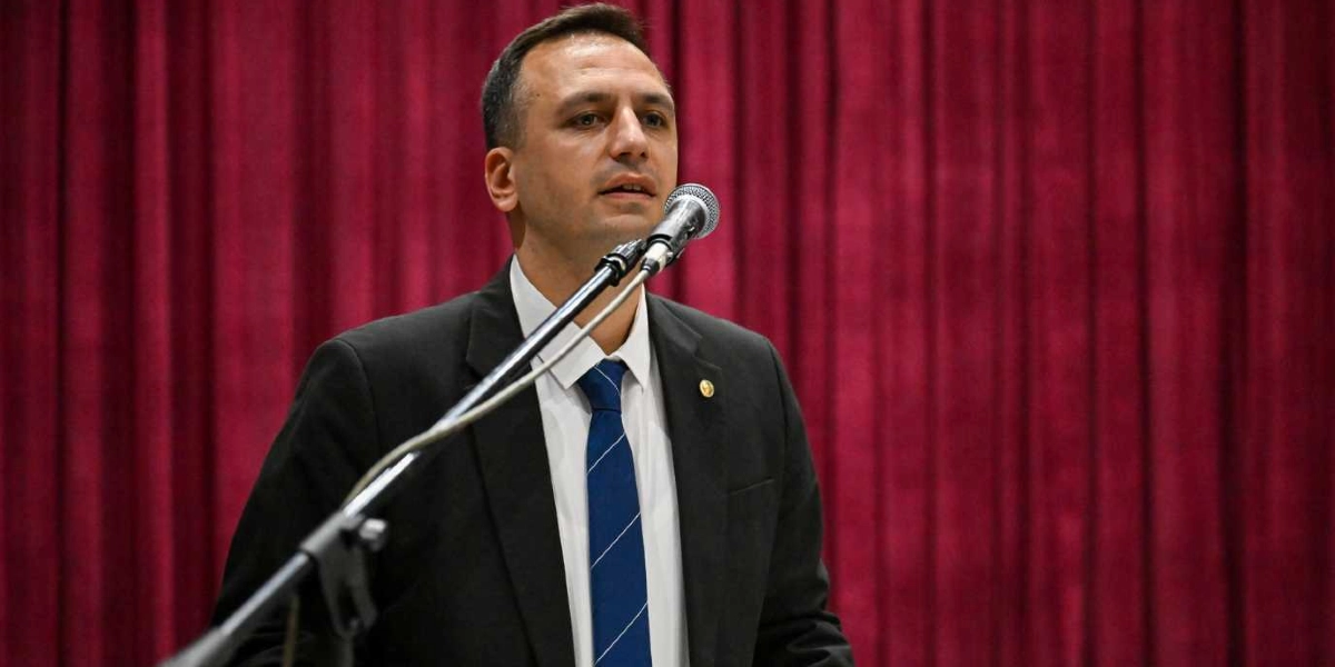 Koyu renk takım elbise ve mavi çizgili kravat giymiş bir erkek, kırmızı perde fonu önünde mikrofon başında konuşma yapıyor.