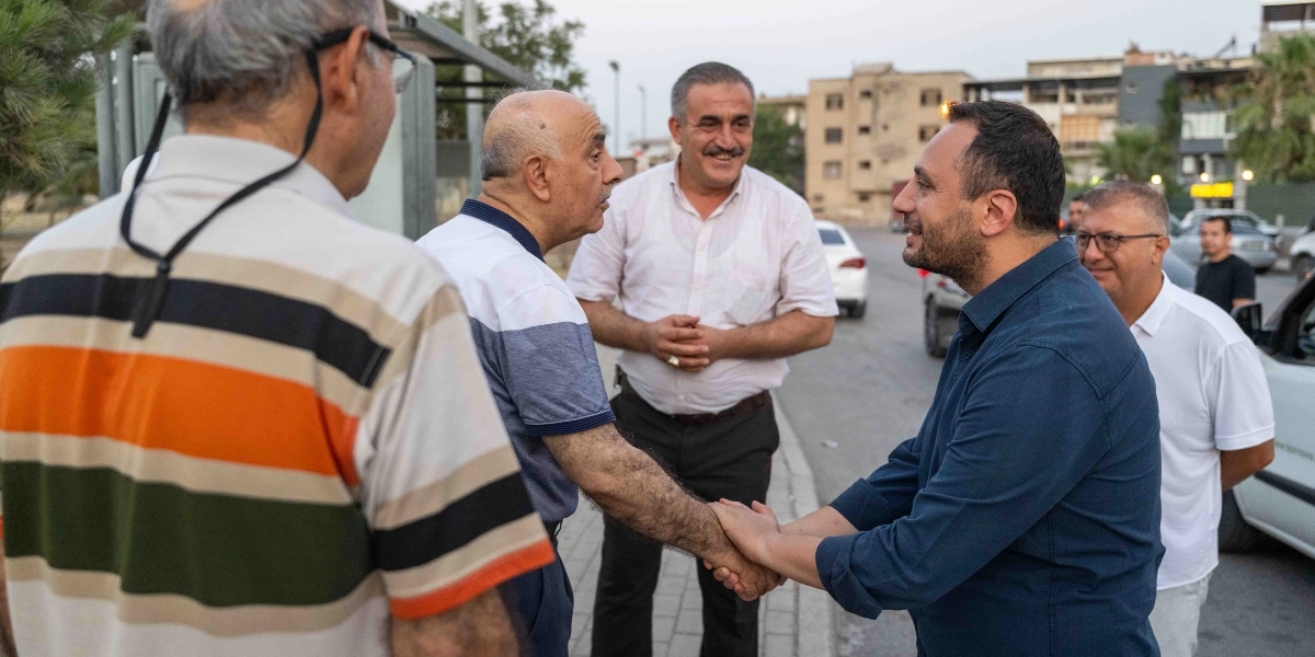 Koyu mavi gömlekli bir erkek, sokakta karşılaştığı yaşlı bir adamın elini tutarak onunla samimi bir şekilde sohbet ediyor. Çevredeki diğer erkekler de gülümseyerek bu diyaloğu izliyor. Arka planda ise binalar ve park halindeki araçların bulunduğu bir mahalle sokağı yer alıyor.