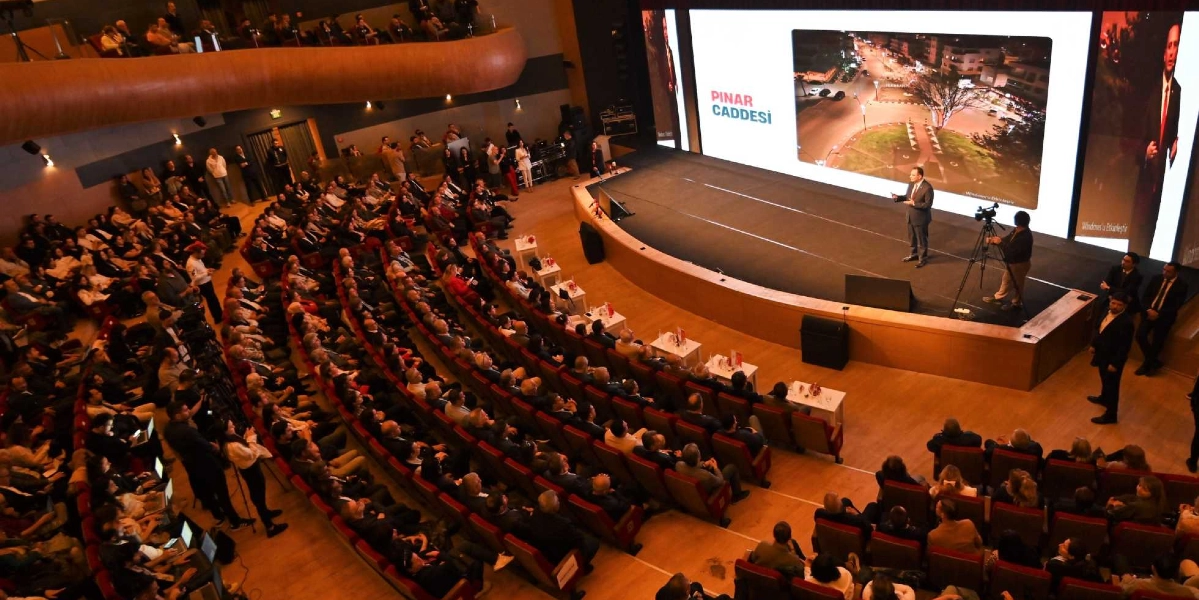 Modern bir konferans salonunda, sahnedeki dev ekranda Bornova Pınar Caddesi'nin gece çekilmiş bir fotoğrafı yansıtılırken, takım elbiseli bir konuşmacı sunum yapıyor. Salon, balkonlu oturma düzeniyle dolu bir izleyici kitlesine ev sahipliği yapıyor; sahnenin sağ tarafında ise bir kameraman çekim yapıyor.