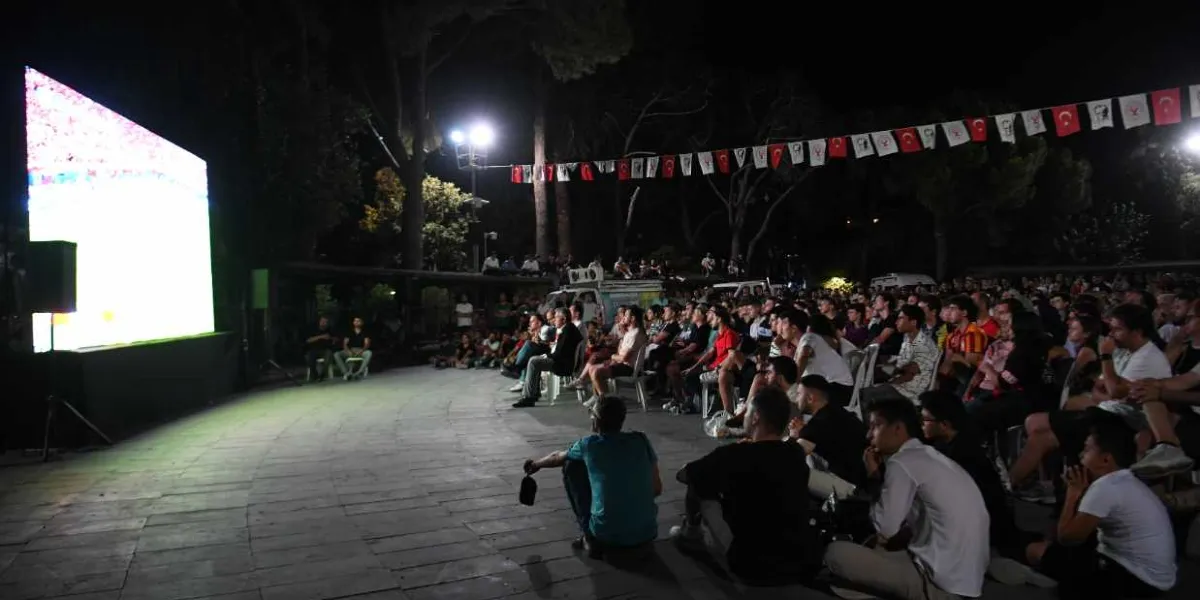 Gece vakti açık havada kurulan dev ekranda basketbol maçı izleyen kalabalık bir grup insan. İnsanlar parke zemin üzerinde sandalyelerde oturarak veya yere bağdaş kurarak maçı takip ediyor. Alanın üzerinde Türk bayrakları ve flamalar asılı duruyor, ortam aydınlatma direkleriyle aydınlatılıyor.