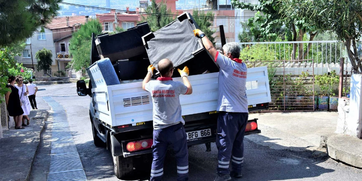 Bornova Belediyesi Temizlik İşleri Müdürlüğü üniformalı iki belediye çalışanı, bir kamyonetin kasasına kullanılamaz haldeki eski mobilyaları ve eşyaları yüklüyor. Sokak üzerinde gerçekleşen bu çalışma sırasında, kaldırımda duran üç kişi de süreci izliyor.
