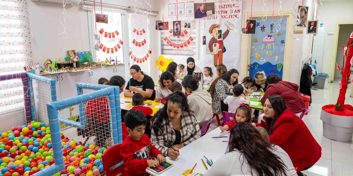 Cumhuriyet Bayramı için süslenmiş bir anaokulu sınıfında, öğretmenleri ve velileriyle birlikte masalarda resim yapan ve boyama etkinliğine katılan çocuklar görülüyor. Sınıfın duvarları Türk bayrakları, Atatürk fotoğrafları ve 