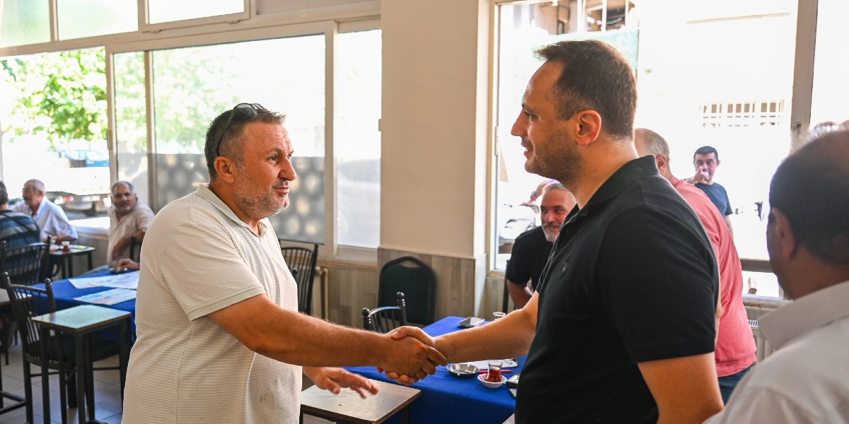 Belediye Başkanı, aydınlık bir kafeteryada beyaz tişörtlü bir vatandaşla tokalaşırken, çevredeki diğer vatandaşlar masalarda oturarak veya ayakta onları izliyor.