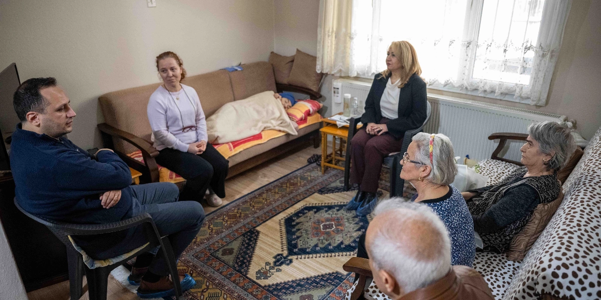 Ev ortamında bir araya gelmiş bir grup insan, odadaki kanepede yatan bir hastayı ziyaret ediyor. Solda lacivert kazaklı bir adam otururken, yanında açık renkli bir kadın bulunuyor. Kanepede yatan kişinin üzeri örtülü ve başı sarılı. Sağ tarafta ise siyah ceketli bir kadın, beyaz saçlı bir kadın ve sırtı dönük oturan yaşlı bir adam yer alıyor. Odanın ortasında desenli bir halı serili ve pencere önünde bir radyatör bulunuyor.