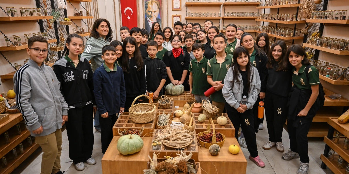 Bir grup ilköğretim öğrencisi ve öğretmenleri, duvarlarında raflar dolusu cam kavanozların dizili olduğu bir dükkan veya atölye ortamında bir arada duruyor. Arka planda Türk bayrağı ve Mustafa Kemal Atatürk portresi asılı. Öğrencilerin önündeki ahşap tezgahlarda ise hasır sepetler, çeşitli kabaklar, kurutulmuş bitkiler ve doğal ürünler sergileniyor. Öğrencilerin çoğu okul forması giyiyor ve hepsi gülümseyerek kameraya bakıyor.