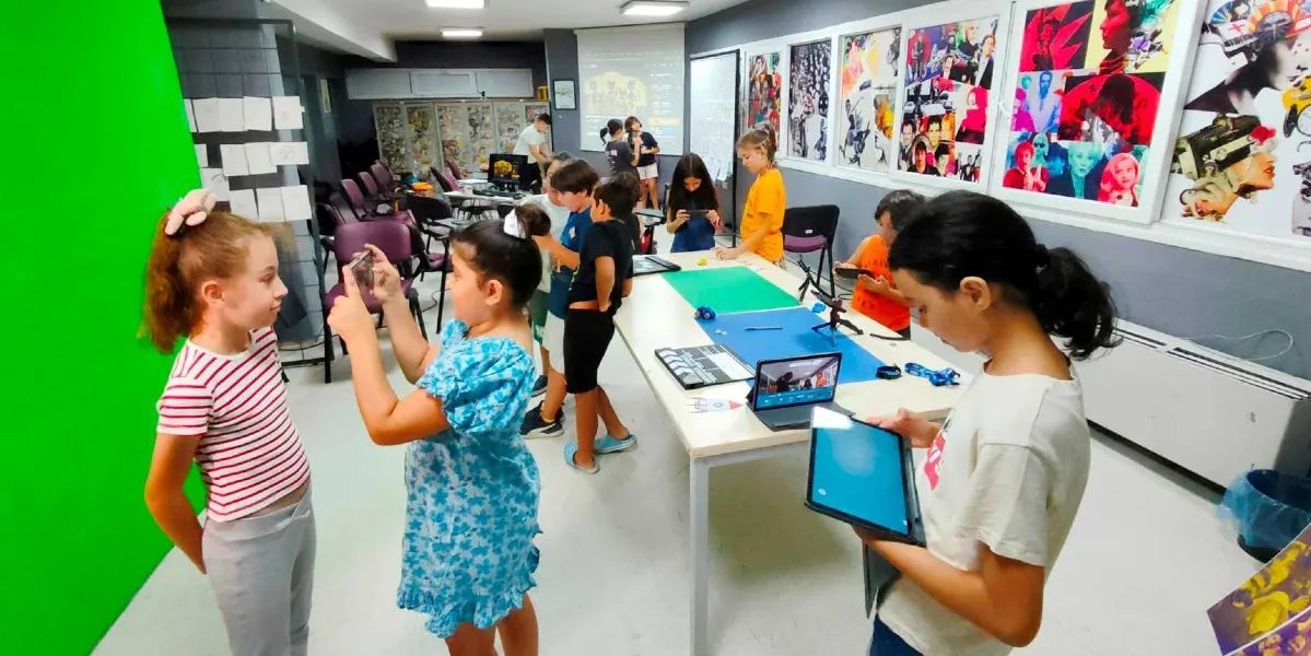 Bir atölye ortamında ellerinde tablet ve telefonlarla çekim yapan veya uygulama kullanan çocuklar. Sol tarafta büyük bir yeşil ekran fonu önünde duran iki kız çocuğu, sağ tarafta ise elinde tablet tutan bir başka kız çocuğu bulunuyor. Ortadaki uzun masanın üzerinde ise bir film çekim klaketi, küçük figürler ve farklı renklerde zeminler yer alıyor. Arka planda duvarlarda sinema temalı renkli posterler ve projeksiyon yansıtılmış bir ekran görülüyor.