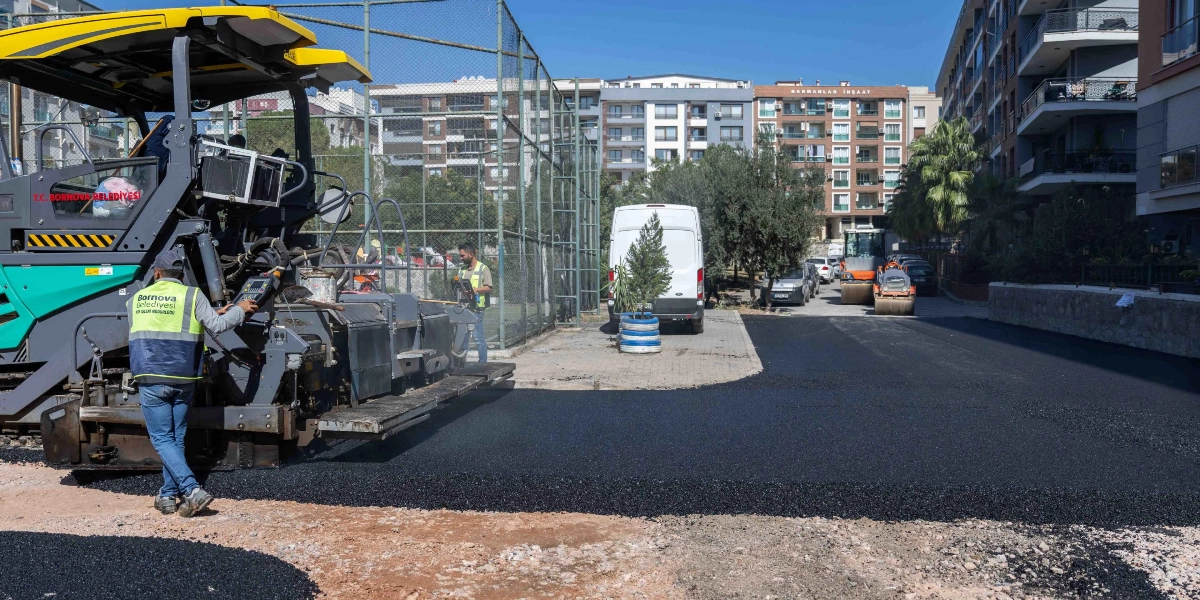 Bornova Belediyesi ekipleri, Erzene mahallesinde bir sokakta asfalt serme makinesi ve silindir kullanarak yol yenileme çalışması yapıyor. Sarı yelekli belediye personeli asfalt serme makinesini yönetirken, arka planda yeni serilmiş asfalt ve çevredeki apartmanlar görülüyor.