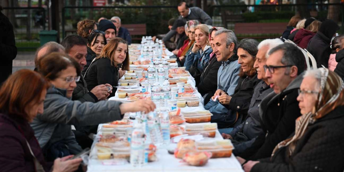 Açık havada kurulan uzun bir masada yan yana oturan kalabalık bir grup insan, iftar yemeği için bir araya gelmiş. Masanın üzerinde plastik kaplarda sunulan yemekler ve su şişeleri dizili durumda. İnsanlar sohbet ederek yemeklerini bekliyor veya yiyorlar; ortamda samimi ve toplu bir iftar buluşması havası hakim.